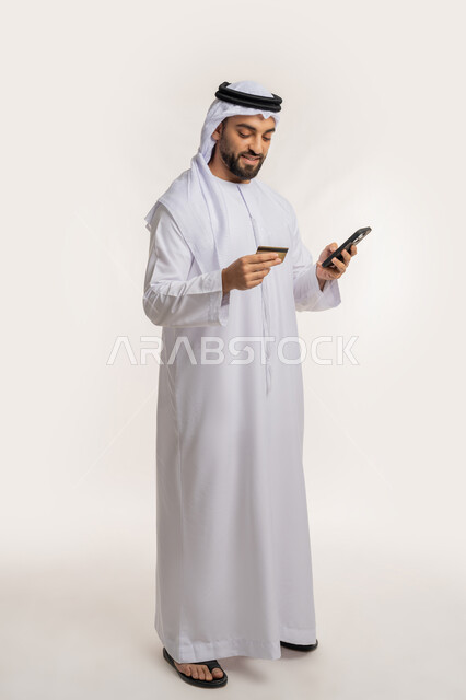 الدفع عبر الانترنت، استخدام جهاز تقني حديث متطور، إمكانية الشراء والدفع من خلال برامج وتطبيقات الجوال، بورتريه لرجل عربي خليجي اماراتي مبتسم يرتدي الكندورة والغترة يحمل بيده الهاتف المحمول وبطاقة الإئتمان البنكية، الجسم بالطول الكامل، خلفية بيضاء