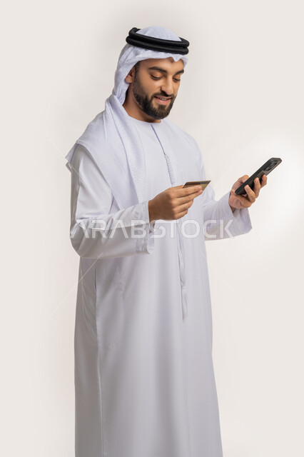 إمكانية الشراء والدفع من خلال برامج وتطبيقات الجوال، مفهوم الدفع عبر الانترنت، استخدام جهاز تقني حديث، بورتريه لرجل عربي خليجي اماراتي مبتسم يرتدي الكندورة والغترة يحمل بيده الهاتف المحمول وبطاقة الإئتمان البنكية، خلفية بيضاء