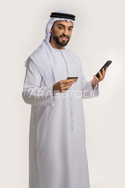 Possibility of purchasing and paying through mobile programs and applications, the concept of online payment, using a modern technical device, portrait of a smiling Emirati Gulf Arab man wearing a kandura and a ghutra holding a mobile phone and a bank credit card, white background