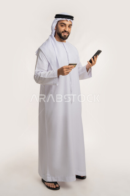 الدفع عبر الانترنت، استخدام جهاز تقني حديث متطور، إمكانية الشراء والدفع من خلال برامج وتطبيقات الجوال، بورتريه لرجل عربي خليجي اماراتي مبتسم يرتدي الكندورة والغترة يحمل بيده الهاتف المحمول وبطاقة الإئتمان البنكية، الجسم بالطول الكامل، خلفية بيضاء