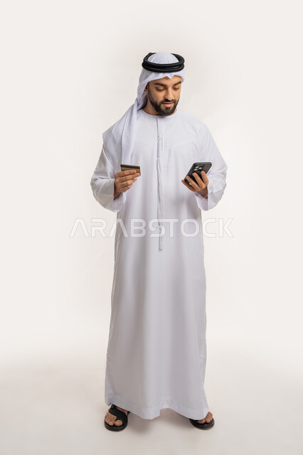 Using a modern advanced technical device, the concept of online payment, the possibility of purchasing and paying through mobile programs and applications, a portrait of a smiling Emirati Gulf Arab man wearing a kandura and a ghutra holding a mobile phone and a bank credit card, full body length, white background