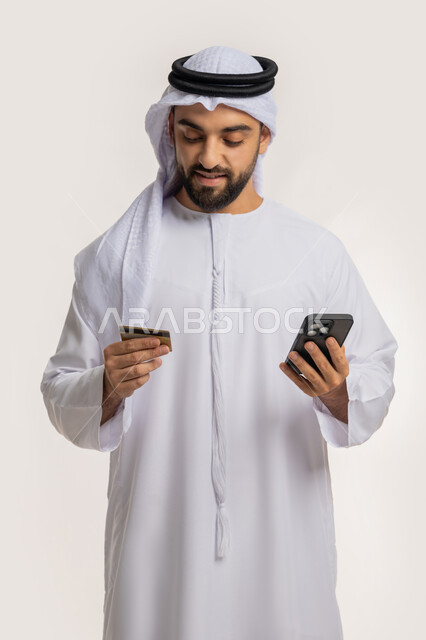 مفهوم الدفع عبر الانترنت، استخدام جهاز تقني حديث، إمكانية الشراء والدفع من خلال برامج وتطبيقات الجوال، بورتريه لرجل عربي خليجي اماراتي مبتسم يرتدي الكندورة والغترة يحمل بيده الهاتف المحمول وبطاقة الإئتمان البنكية، خلفية بيضاء