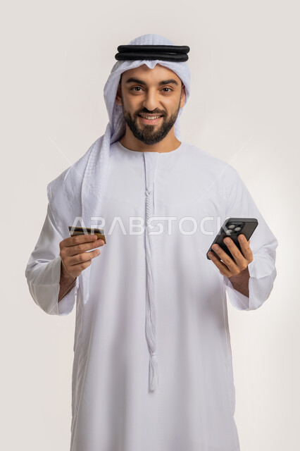 مفهوم الدفع عبر الانترنت، استخدام جهاز تقني حديث، إمكانية الشراء والدفع من خلال برامج وتطبيقات الجوال، بورتريه لرجل عربي خليجي اماراتي مبتسم يرتدي الكندورة والغترة يحمل بيده الهاتف المحمول وبطاقة الإئتمان البنكية، خلفية بيضاء