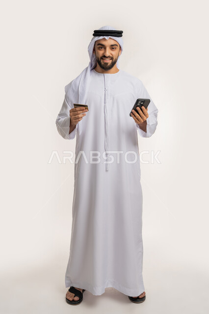 استخدام جهاز تقني حديث، مفهوم الدفع عبر الانترنت، إمكانية الشراء والدفع من خلال برامج وتطبيقات الجوال، بورتريه لرجل عربي خليجي اماراتي مبتسم يرتدي الكندورة والغترة يحمل بيده الهاتف المحمول وبطاقة الإئتمان البنكية، الجسم بالطول الكامل، خلفية بيضاء