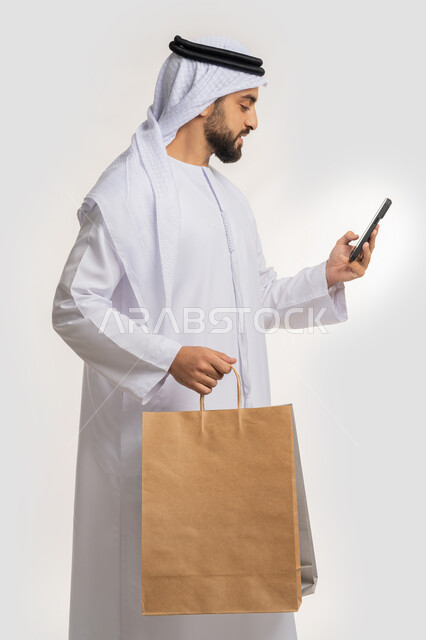 استخدام التكنولوجيا في الحياة اليومية، التسوق وشراء المستلزمات والهدايا عبر الانترنت، ايماءات وتعابير السرور والسعادة، بورتريه من الجانب لشاب عربي خليجي اماراتي يرتدي الغترة والكندورة يمسك بيده الهاتف المحمول وأكياس التسوق، خلفية بيضاء