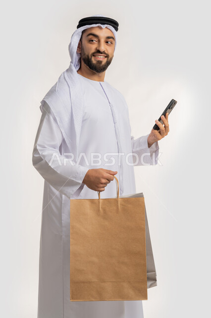 ايماءات وتعابير السرور والسعادة، استخدام التكنولوجيا في الحياة اليومية، التسوق وشراء المستلزمات والهدايا عبر الانترنت، بورتريه لشاب عربي خليجي اماراتي يرتدي الغترة والكندورة يمسك بيده الهاتف المحمول وأكياس التسوق، خلفية بيضاء