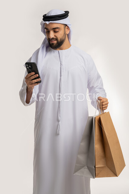 ايماءات وتعابير السرور والسعادة، استخدام التكنولوجيا في الحياة اليومية، التسوق وشراء المستلزمات والهدايا عبر الانترنت، بورتريه لشاب عربي خليجي اماراتي يرتدي الغترة والكندورة يمسك بيده الهاتف المحمول وأكياس التسوق، خلفية بيضاء