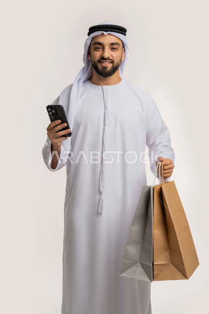 ايماءات وتعابير السرور والسعادة، استخدام التكنولوجيا في الحياة اليومية، التسوق وشراء المستلزمات والهدايا عبر الانترنت، بورتريه لشاب عربي خليجي اماراتي يرتدي الغترة والكندورة يمسك بيده الهاتف المحمول وأكياس التسوق، خلفية بيضاء