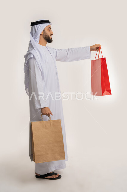 الاستفادة من العروض والتخفيضات الموسمية، المتعة في التسوق واقتناء المستلزمات والهدايا، بورتريه من الجانب لرجل عربي خليجي اماراتي مبتسم يرتدي الكندورة والغترة يقدم أكياس ورقية ملونة بإيماءات السرور والسعادة، صورة الجسم بالطول الكامل، خلفية بيضاء