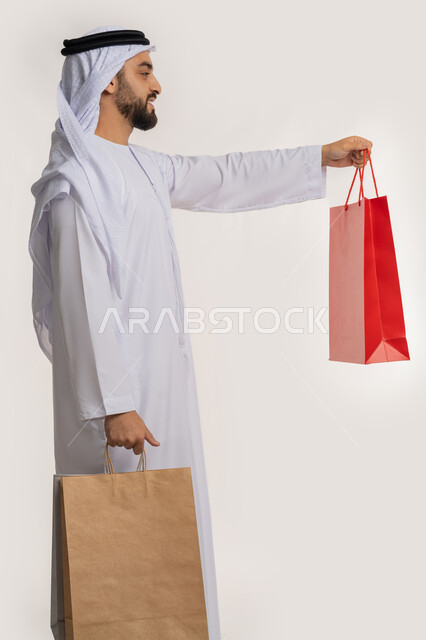 الاستفادة من العروض والتخفيضات الموسمية، المتعة في التسوق واقتناء المستلزمات والهدايا، بورتريه من الجانب لرجل عربي خليجي اماراتي مبتسم يرتدي الكندورة والغترة يقدم أكياس ورقية ملونة بإيماءات السرور والسعادة، خلفية بيضاء