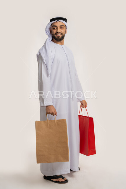 الاستفادة من العرض والخصم في المول، اقتناء الهدايا والحاجات الضرورية، النظر الى الكاميرا بايماءات السعادة، قضاء وقت ممتع في شراء المستلزمات، بورتريه لشاب عربي خليجي اماراتي مبتسم يرتدي الكندورة والغترة يحمل بيده أكياس التسوق الملونة، الجسم بالطول الكامل، خلفية بيضاء