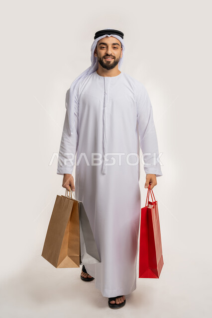 الاستفادة من العرض والخصم في المول، اقتناء الهدايا والحاجات الضرورية، النظر الى الكاميرا بايماءات السعادة، قضاء وقت ممتع في شراء المستلزمات، بورتريه لشاب عربي خليجي اماراتي مبتسم يرتدي الكندورة والغترة يحمل بيده أكياس التسوق الملونة، الجسم بالطول الكامل، خلفية بيضاء