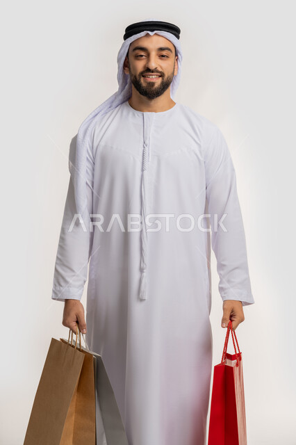 النظر الى الكاميرا بايماءات السعادة والسرور، اقتناء الهدايا والحاجات الضرورية، الاستفادة من العرض والخصم في المول، قضاء وقت ممتع في شراء المستلزمات، بورتريه لشاب عربي خليجي اماراتي مبتسم يرتدي الكندورة والغترة يحمل بيده أكياس التسوق الملونة، خلفية بيضاء