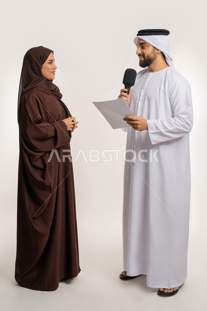 إجراء المقابلة وطرح الأسئلة، بورتريه لرجل عربي خليجي إماراتي يرتدي الكندورة والغترة يحمل بيده مكبر الصوت وورقة يقوم بمناقشة إمرأة ترتدي الحجاب والعباءة، التواصل الفعال وتبادل التساؤولات، صورة الجسم بالطول الكامل، خلفية بيضاء