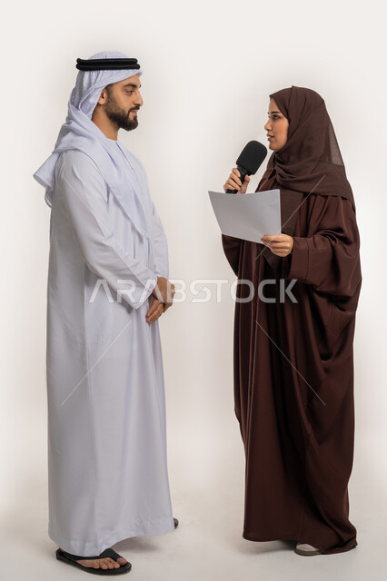 التواصل الفعال وتبادل التساؤولات، إجراء المقابلة وطرح الأسئلة، بورتريه لإمرأة عربية خليجية إماراتية ترتدي الحجاب والعباءة البنية تحمل بيدها مكبر الصوت وورقة تقوم بمناقشة رجل يرتدي الكندورة والغترة، صورة الجسم بالطول الكامل، خلفية بيضاء