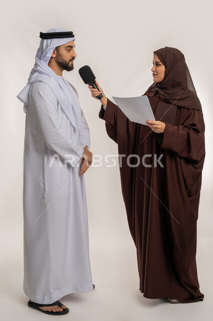 التواصل الفعال وتبادل التساؤولات، إجراء المقابلة وطرح الأسئلة، بورتريه لإمرأة عربية خليجية إماراتية ترتدي الحجاب والعباءة البنية تحمل بيدها مكبر الصوت وورقة تقوم بمناقشة رجل يرتدي الكندورة والغترة، صورة الجسم بالطول الكامل، خلفية بيضاء
