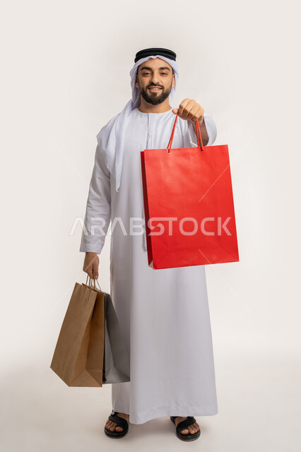 اقتناء الهدايا والحاجات الضرورية، النظر الى الكاميرا بايماءات السعادة، الاستفادة من العرض والخصم في المول، قضاء وقت ممتع في شراء المستلزمات، بورتريه لشاب عربي خليجي اماراتي مبتسم يرتدي الكندورة والغترة يحمل بيده أكياس التسوق الملونة، الجسم بالطول الكامل، خلفية بيضاء