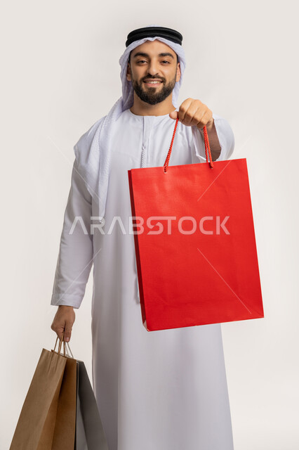 النظر الى الكاميرا بايماءات السعادة والسرور، اقتناء الهدايا والحاجات الضرورية، الاستفادة من العرض والخصم في المول، قضاء وقت ممتع في شراء المستلزمات، بورتريه لشاب عربي خليجي اماراتي مبتسم يرتدي الكندورة والغترة يحمل بيده أكياس التسوق الملونة، خلفية بيضاء