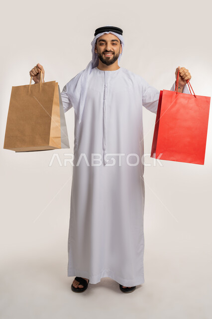 اقتناء الهدايا والحاجات الضرورية، النظر الى الكاميرا بايماءات السعادة، الاستفادة من العرض والخصم في المول، قضاء وقت ممتع في شراء المستلزمات، بورتريه لشاب عربي خليجي اماراتي مبتسم يرتدي الكندورة والغترة يحمل بيده أكياس التسوق الملونة، الجسم بالطول الكامل، خلفية بيضاء