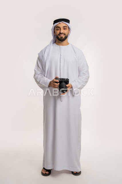 ايماءات وتعابير السرور والسعادة، احتراف التصوير الفوتوغرافي باستخدام الكاميرات الرقمية, ممارسة هواية التصوير, بورتريه لشاب عربي خليجي اماراتي مبتسم يرتدي الكندورة والغترة يقوم بالتقاط صور تذكارية عبر كاميرا فوتوغرافية, الجسم بالطول الكامل، خلفية بيضاء