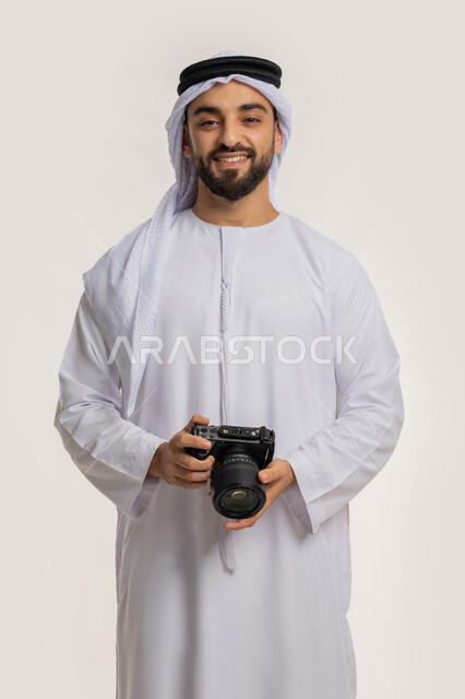 ايماءات وتعابير السرور والسعادة، احتراف التصوير الفوتوغرافي باستخدام الكاميرات الرقمية, ممارسة هواية التصوير, بورتريه من الجانب لشاب عربي خليجي اماراتي مبتسم يرتدي الكندورة والغترة يقوم بالتقاط صور تذكارية عبر كاميرا فوتوغرافية, خلفية بيضاء
