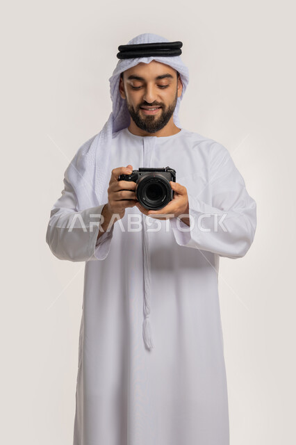 Professional photography using digital cameras, professional photographer reviewing his shots, practicing photography hobby, portrait of a smiling young Arab Gulf Emirati man wearing a kandura and ghutra taking souvenir photos using a photographic camera, white background