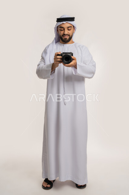 مصور محترف يقوم بمراجعة لقطاته, اتقان التصوير الفوتوغرافي باستخدام الكاميرات الرقمية, ممارسة هواية التصوير, بورتريه لشاب عربي خليجي اماراتي يرتدي الكندورة والغترة يقوم بالتقاط صور تذكارية عبر كاميرا فوتوغرافية, الجسم بالطول الكامل، خلفية بيضاء