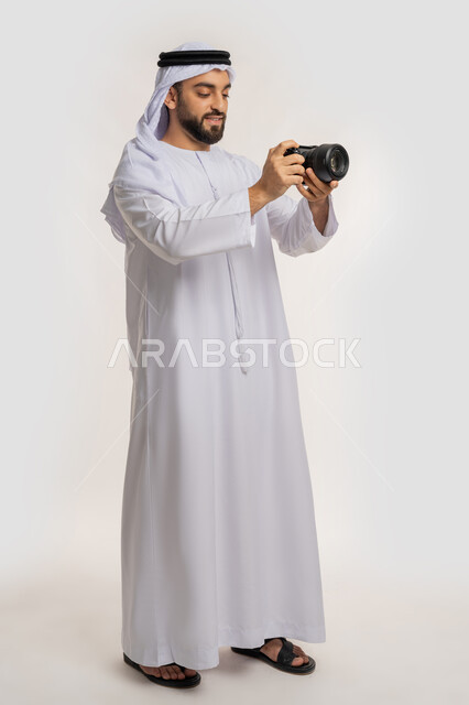 مصور محترف يقوم بمراجعة لقطاته, اتقان التصوير الفوتوغرافي باستخدام الكاميرات الرقمية, ممارسة هواية التصوير, بورتريه من الجانب لشاب عربي خليجي اماراتي يرتدي الكندورة والغترة يقوم بالتقاط صور تذكارية عبر كاميرا فوتوغرافية, الجسم بالطول الكامل، خلفية بيضاء