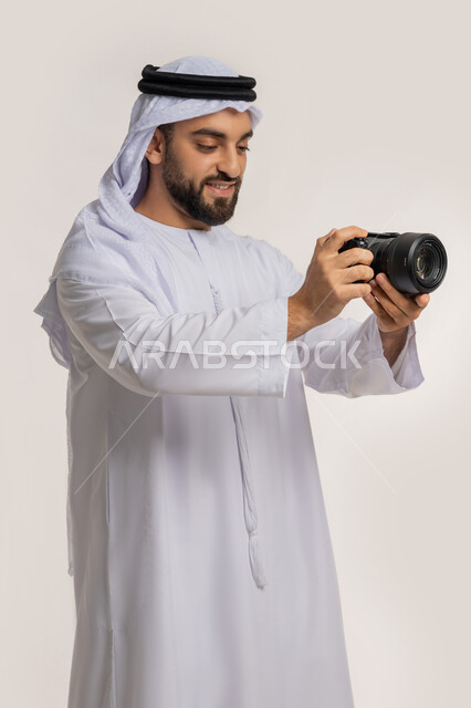 احتراف التصوير الفوتوغرافي باستخدام الكاميرات الرقمية, مصور محترف يقوم بمراجعة لقطاته, ممارسة هواية التصوير, بورتريه لشاب عربي خليجي اماراتي مبتسم يرتدي الكندورة والغترة يقوم بالتقاط صور تذكارية عبر كاميرا فوتوغرافية, خلفية بيضاء