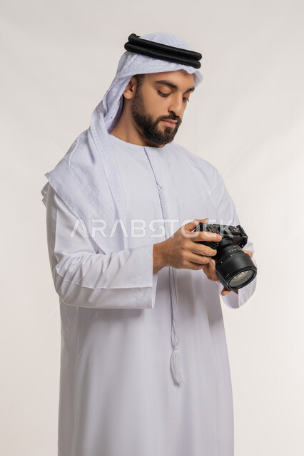 احتراف التصوير الفوتوغرافي باستخدام الكاميرات الرقمية, مصور محترف يقوم بمراجعة لقطاته, ممارسة هواية التصوير, بورتريه لشاب عربي خليجي اماراتي مبتسم يرتدي الكندورة والغترة يقوم بالتقاط صور تذكارية عبر كاميرا فوتوغرافية, خلفية بيضاء