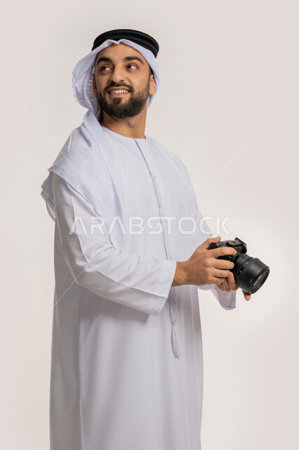 Looking up with expressions of pleasure, professional photography using digital cameras, practicing photography hobby, side portrait of a smiling young Arab Gulf Emirati man wearing a kandura and ghutra taking souvenir photos with a camera, white background