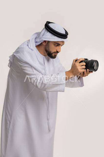 Practicing the hobby of photography with expressions of pleasure, a side portrait of a young Arab Gulf Emirati man wearing a kandura and a ghutra holding a camera in his hand, mastering photography using high-resolution digital cameras, a professional photographer taking souvenir photos, white background