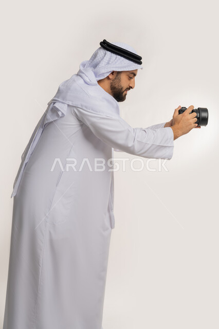 ممارسة هواية التصوير بتعابير السرور, بورتريه من الجانب لشاب عربي خليجي اماراتي يرتدي الكندورة والغترة يمسك بيده كاميرا فوتوغرافية, اتقان التصوير الفوتوغرافي باستخدام كاميرات رقمية عالية الدقة, مصور محترف يقوم بالتقاط صور تذكارية, خلفية بيضاء