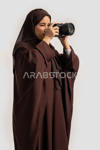 مصورة سعودية تمارس هواية التصوير بتعابير السرور، وظيفة ومهنة نسائية, اتقان فن التصوير الفوتوغرافي باستخدام كاميرات رقمية عالية الدقة, بورتريه لإمراة عربية خليجية اماراتية ترتدي الحجاب والعباءة تقوم بالتقاط صورة تذكارية عبر كاميرا فوتوغرافية, خلفية بيضاء
