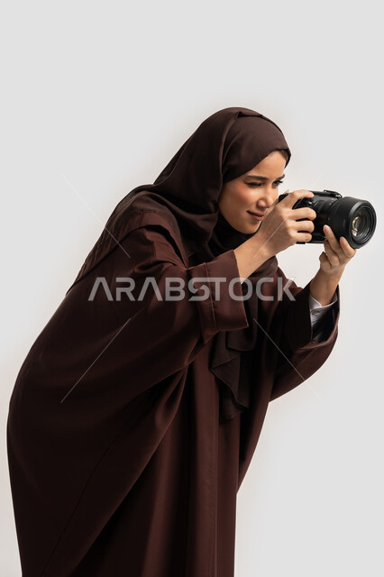 وظيفة ومهنة نسائية, اتقان فن التصوير الفوتوغرافي باستخدام كاميرات رقمية عالية الدقة, بورتريه لإمراة عربية خليجية اماراتية ترتدي الحجاب والعباءة تقوم بالتقاط صورة تذكارية عبر كاميرا فوتوغرافية, مصورة سعودية تمارس هواية التصوير بتعابير السرور، خلفية بيضاء