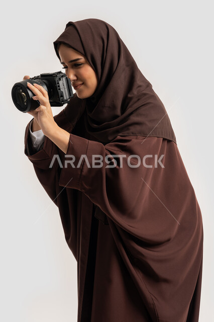 وظيفة ومهنة نسائية, اتقان فن التصوير الفوتوغرافي باستخدام كاميرات رقمية عالية الدقة, بورتريه لإمراة عربية خليجية اماراتية ترتدي الحجاب والعباءة تقوم بالتقاط صورة تذكارية عبر كاميرا فوتوغرافية, مصورة سعودية تمارس هواية التصوير بتعابير السرور، خلفية بيضاء