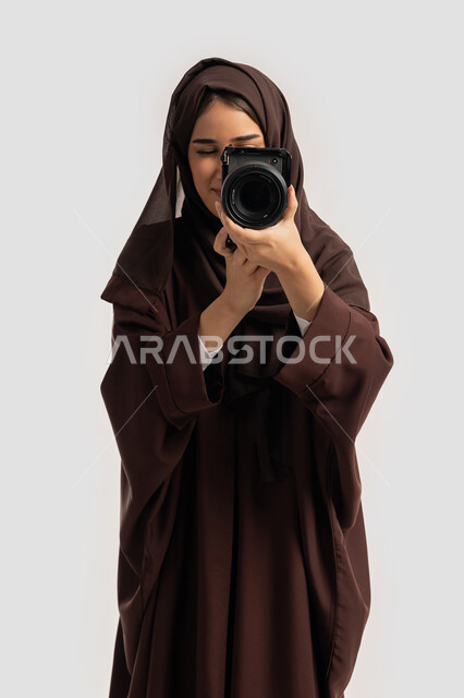 وظيفة ومهنة نسائية, اتقان فن التصوير الفوتوغرافي باستخدام كاميرات رقمية عالية الدقة, بورتريه لإمراة عربية خليجية اماراتية ترتدي الحجاب والعباءة تقوم بالتقاط صورة تذكارية عبر كاميرا فوتوغرافية, مصورة سعودية تمارس هواية التصوير بتعابير السرور، خلفية بيضاء