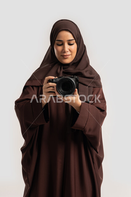 احتراف فن التصوير الفوتوغرافي باستخدام كاميرات رقمية عالية الدقة, وظيفة ومهنة نسائية, بورتريه لإمراة عربية خليجية اماراتية ترتدي الحجاب والعباءة تقوم بالتقاط صورة تذكارية عبر كاميرا فوتوغرافية, مصورة سعودية تمارس هواية التصوير بتعابير السرور، خلفية بيضاء