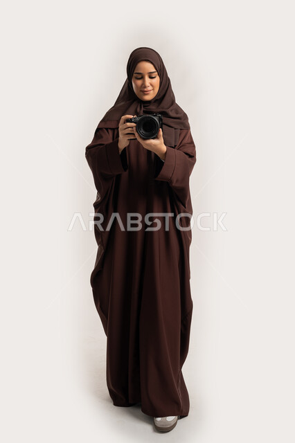 اتقان التصوير الفوتوغرافي باستخدام كاميرات رقمية عالية الدقة, بورتريه لإمراة عربية خليجية اماراتية ترتدي الحجاب والعباءة تقوم بالتقاط صورة تذكارية عبر كاميرا فوتوغرافية, مصورة سعودية تمارس هواية التصوير، وظيفة ومهنة نسائية, الجسم بالطول الكامل، خلفية بيضاء