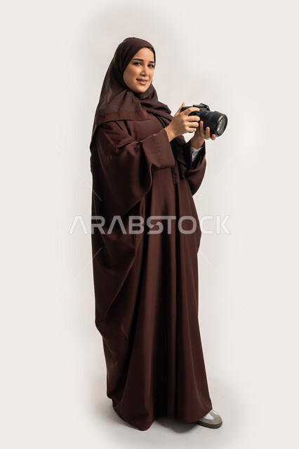 Mastering photography using high-resolution digital cameras, portrait of an Arab Gulf Emirati woman wearing a hijab and abaya taking a souvenir photo with a camera, a Saudi photographer practicing photography as a hobby, a female job and profession, full-length body, white background