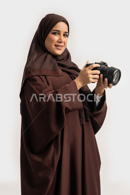 احتراف فن التصوير الفوتوغرافي باستخدام كاميرات رقمية عالية الدقة, وظيفة ومهنة نسائية, بورتريه لإمراة عربية خليجية اماراتية ترتدي الحجاب والعباءة تقوم بالتقاط صورة تذكارية عبر كاميرا فوتوغرافية, مصورة سعودية تمارس هواية التصوير بتعابير السرور، خلفية بيضاء
