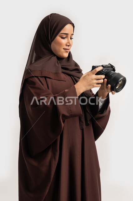 وظيفة ومهنة نسائية, اتقان فن التصوير الفوتوغرافي باستخدام كاميرات رقمية عالية الدقة, بورتريه لإمراة عربية خليجية اماراتية ترتدي الحجاب والعباءة تقوم بالتقاط صورة تذكارية عبر كاميرا فوتوغرافية, مصورة سعودية تمارس هواية التصوير بتعابير السرور، خلفية بيضاء