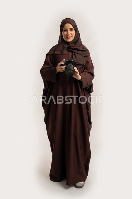 اتقان التصوير الفوتوغرافي باستخدام كاميرات رقمية عالية الدقة, بورتريه لإمراة عربية خليجية اماراتية ترتدي الحجاب والعباءة تقوم بالتقاط صورة تذكارية عبر كاميرا فوتوغرافية, مصورة سعودية تمارس هواية التصوير، وظيفة ومهنة نسائية, الجسم بالطول الكامل، خلفية بيضاء