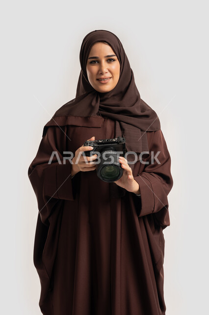 احتراف فن التصوير الفوتوغرافي باستخدام كاميرات رقمية عالية الدقة, وظيفة ومهنة نسائية, بورتريه لإمراة عربية خليجية اماراتية ترتدي الحجاب والعباءة تقوم بالتقاط صورة تذكارية عبر كاميرا فوتوغرافية, مصورة سعودية تمارس هواية التصوير بتعابير السرور، خلفية بيضاء