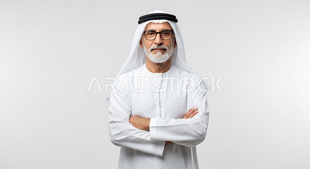 مفهوم الأناقة والرجولة، بورتريه لرجل عربي خليجي اماراتي يرتدي الكندورة والغترة يقف بأيدي متقاطعة ينظر إلى الكاميرا بإيماءات الجدية والثقة بالنفس، الاهتمام بالمظهر الخارجي، خلفية بيضاء