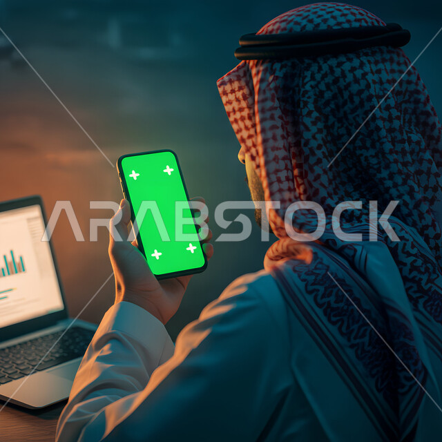 العمل على الحاسوب المحمول، دمج التكنولوجيا الحديثة بالحياة العملية، عرض شاشة خضراء فارغة، استخدام جهاز تقني حديث، صورة مقربة من الجانب لرجل عربي خليجي سعودي يرتدي الثوب التقليدي والشماغ يمسك الهاتف المحمول، تصفح مواقع التواصل الاجتماعي عبر الجوال، كروما