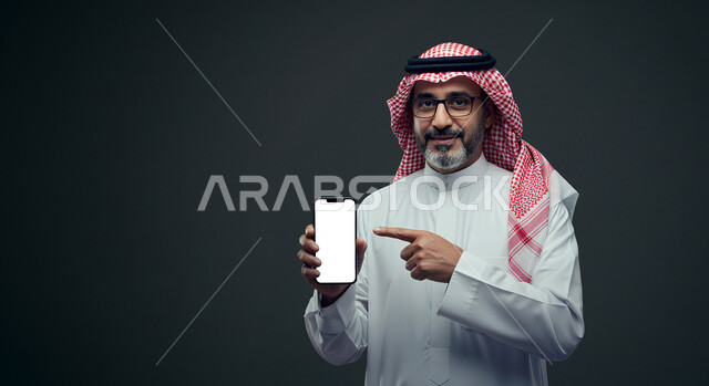 استخدام جهاز تقني حديث، الاشارة باصبع السبابة إلى شاشة بيضاء فارغة، بورتريه مقرب لرجل عربي خليجي سعودي يرتدي الثوب التقليدي والشماغ والنظارة الطبية يمسك بيده الهاتف المحمول ينظر إلى الكاميرا بإيماءات السرور، تصفح مواقع التواصل الاجتماعي عبر الجوال، خلفية رمادية
