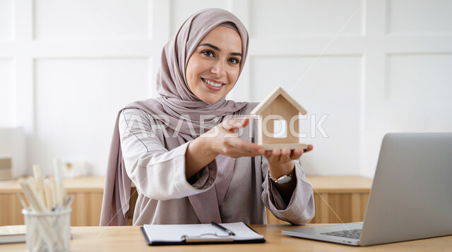 استخدام جهاز تقني حديث، الاسكان والتمليك اونلاين، بيع وشراء العقارات عبر الانترنت، امراة عربية خليجية سعودية ترتدي الحجاب تعمل على الحاسوب المحمول تحمل بيدها مجسم خشبي لمنزل صغير تنظر إلى الكاميرا بإيماءات السرور، مهنة ووظيفة مكتبية