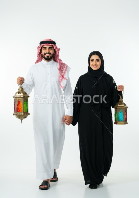 الاحتفال بقدوم شهر الخير، الاستعداد لاستقبال شهر رمضان المبارك، بورتريه لرجل عربي خليجي سعودي يرتدي الشماغ والثوب التقليدي يمشي برفقة زوجته يحملان فانوس معدني مزخرف، زينة وأنوار الأعياد والمناسبات، الجو الرمضاني الروحاني، الجسم بالطول الكامل، خلفية بيضاء 
