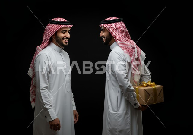 مفهوم الشكر والامتنان، تقديم الهدايا بالاعياد والمناسبات الخاصة، التعبير عن الحب والتقدير والاحترام، بورتريه لرجل عربي سعودي يرتدي الثوب التقليدي والشماغ يحمل بيديه هدية يخبئها خلف ظهره يستعد لتقديمها إلى أخيه، المحبة بين الأصدقاء، خلفية سوداء
